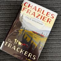 Book cover for The Trackers by Charles Frazier. Cover features a drawing of a landscape with mountains, trees, and winding roads.