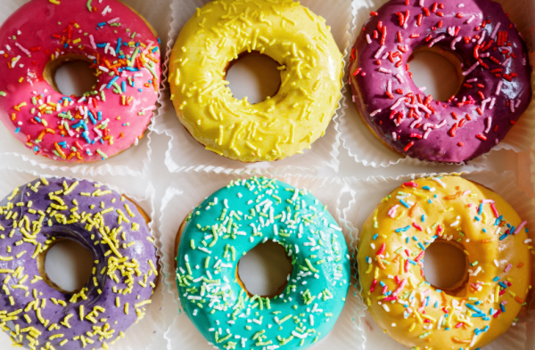 Six donuts in blue, pink, purple, yellow frosting with multicolored sprinkles on a light background.
