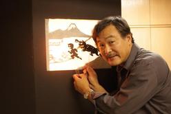 Man sits in front of shadow puppet stage. On the stage a monster is chasing a running person with a mountain in the background.