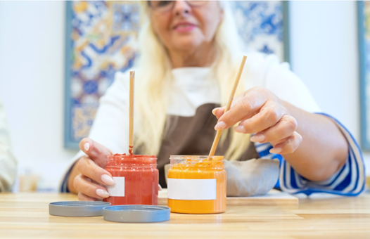 A woman using paint brushes in two jars of paint - one orange and one yellow.