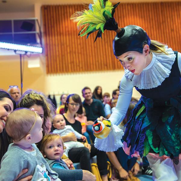 Opera performer wearing bird costume shows a toy bird to a baby and audience of kids and parents