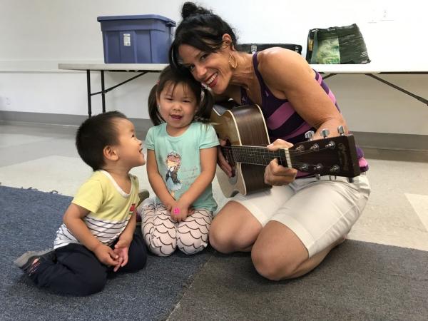 A woman holding a guitar kneeling on the floor with two young children.
