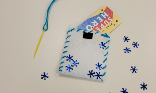 A white rectangle of felt sewed up three sides with blue thread, yellow needle still attached. The pouch is decorated with blue, snowflake-shaped sequins and has a gift card sticking out of it.