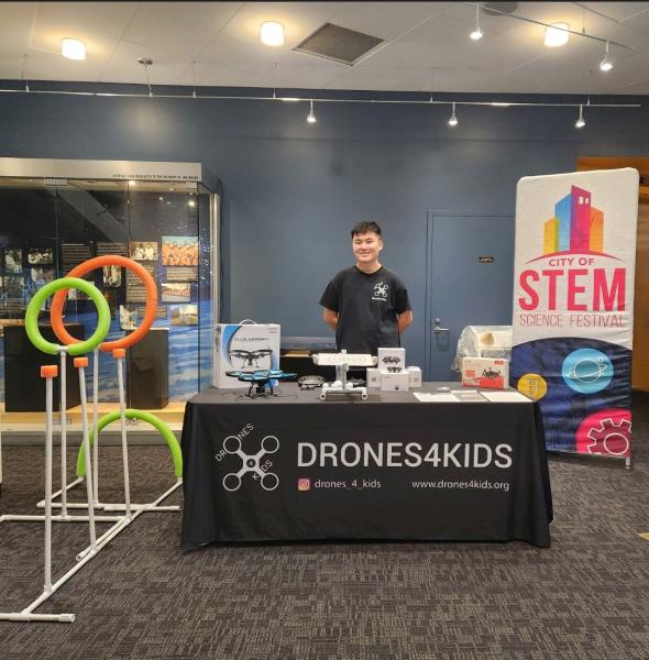 Drone4Kids setup, in center a gentleman stands behind a table with company name. To right is vertical STEM banner, to left are hoops and circles that drones will fly through.