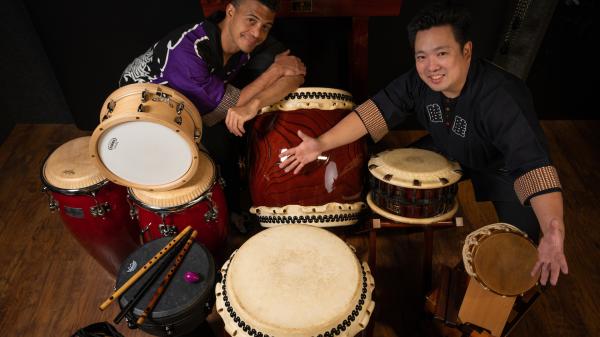 Two men smiling  with many drums surrounding them 