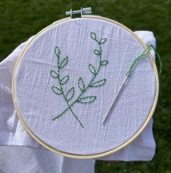 A white cloth in an embroidery hoop with two leafy branches embroidered on it in green thread. A needle with more green thread rests on top of the fabric.