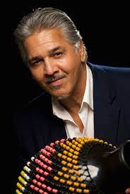 A Latino man with facial hair in a blue suit holds a percussion instrument built out of a hallow base wrapped in colorful beads. 