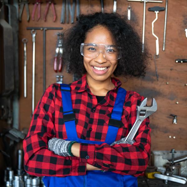 Teenage girl wearing safety goggles and gloves and holding wrench in front of wall of tools