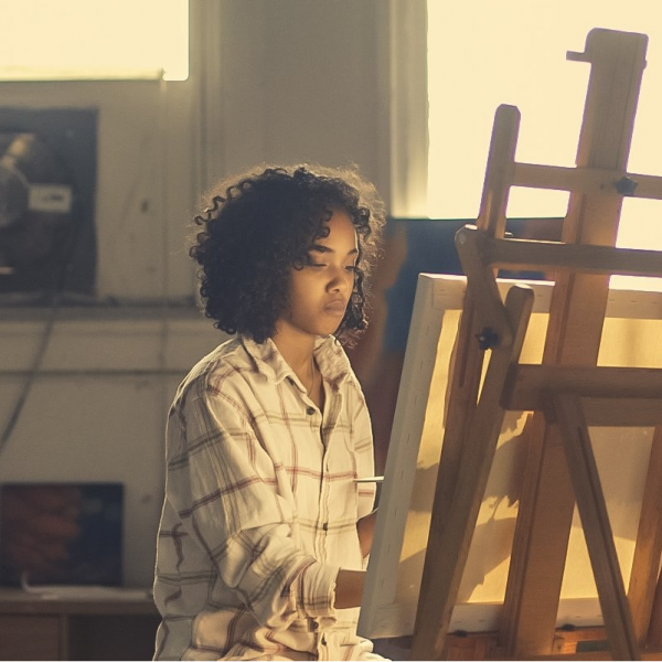 Teen sitting at easel in front of canvas focused on painting.