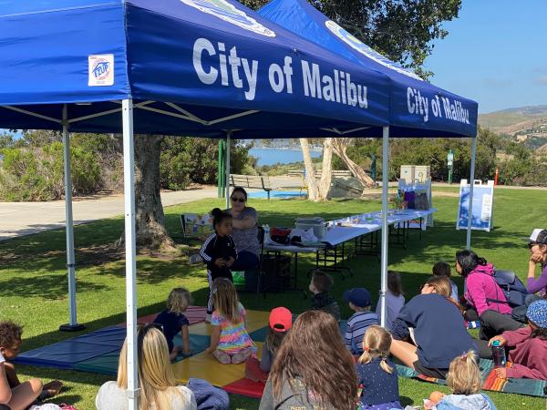 A crowd of families sits on the grass in front of blue "City of Malibu" tents while a librarian reads a book to them.