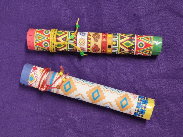 Two rain stick tubes covered in brightly-patterned paper and tied with twine on a purple background.