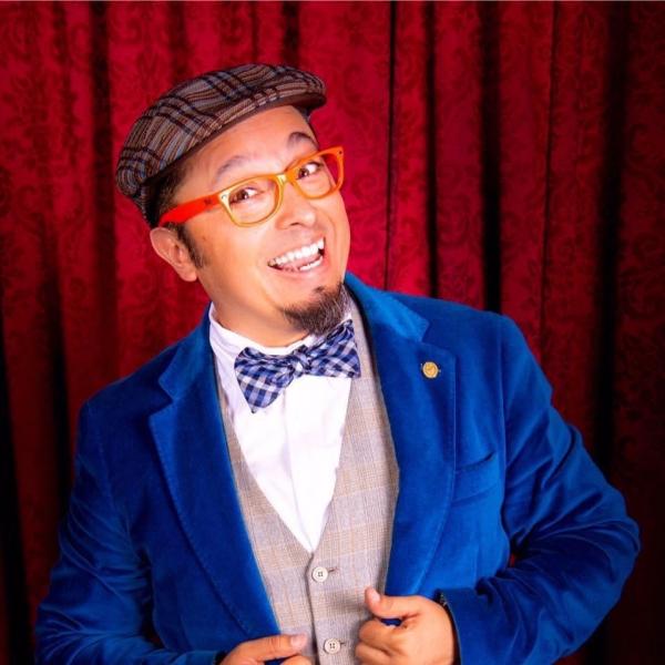 Wacko is smiling at the camera wearing a blue jacket and blue bow tie over a grey vest and white shirt. He is wearing orange rimmed glasses and a plaid page boy hat.