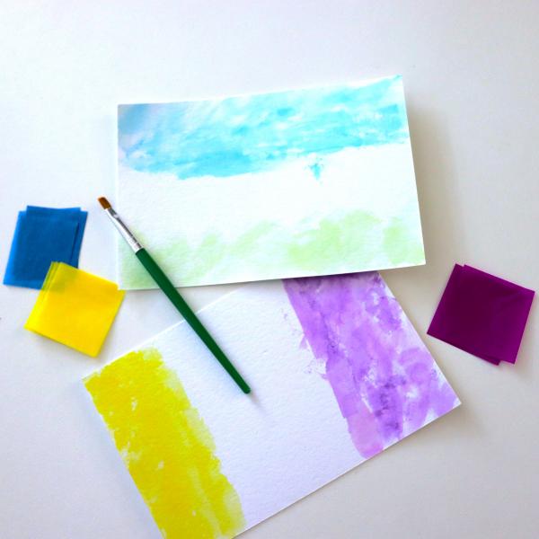White postcards painted with stripes of color surrounded by tissue paper squares and a paint brush.
