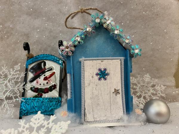 A blue house with white door made from craft supplies sitting next to a miniature sled and round silver ornament.