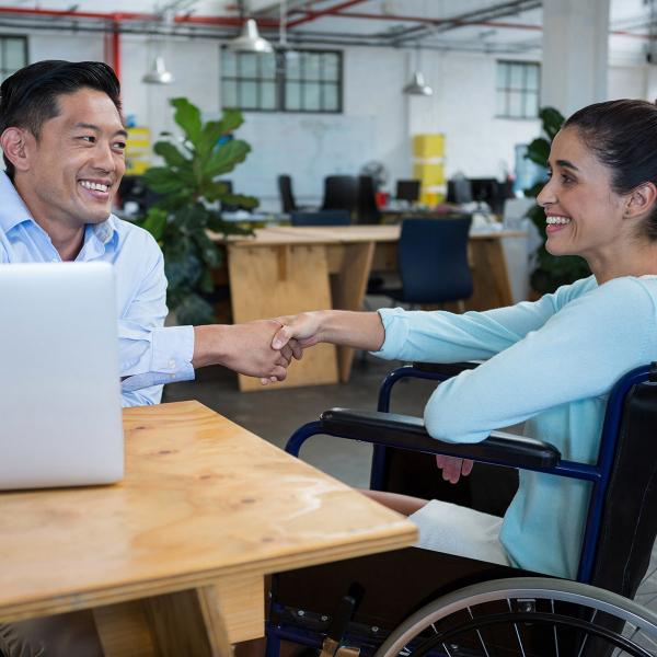 Man in front of laptop shaking hands with woman in wheelchair in office setting.
