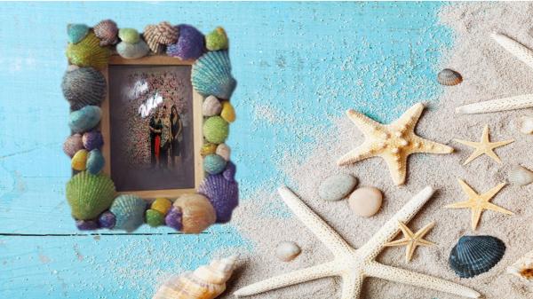 Photo frame covered by colorful seashells next to rocks, starfish, and seashells laying on sand and blue wood.
