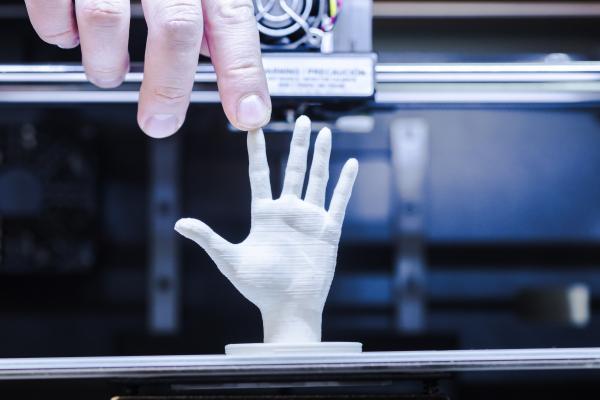 Image of a person's finger touching a miniature white 3D-printed model of a hand on a 3d printer bed