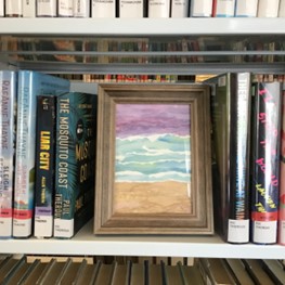 Photo of framed ocean landscape painting on a book shelf between library books.