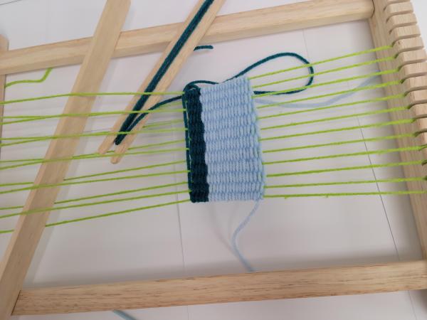Wooden loom with green warp threads and blue weft threads part way up. 
