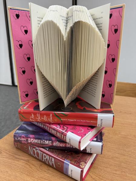 An open book with pages folded in a shape of a heart a top of four red & pink colored books on a wooden table