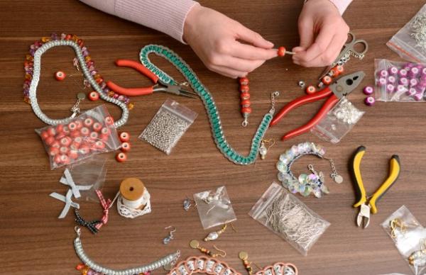Hands stringing beads on a wire. On the table are various beads, cords, and tools.