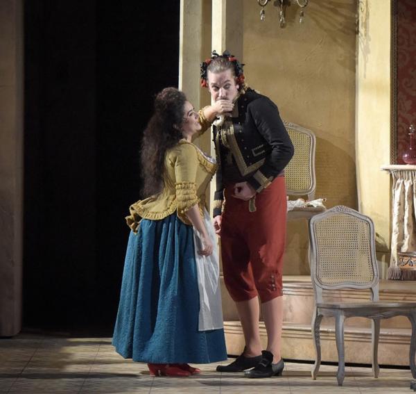 Two opera singers in historical costume, the woman is putting her hand over the man's mouth
