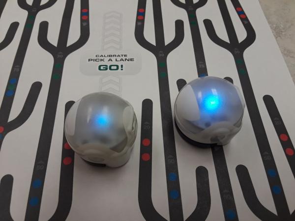 Two spherical Ozobots with blue lights glowing inside on a paper with black lines drawn on it.