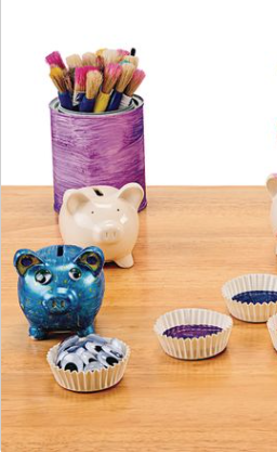 Two piggy banks, one painted blue, the other unpainted. There are paint brushes and googly eyes next to them. 